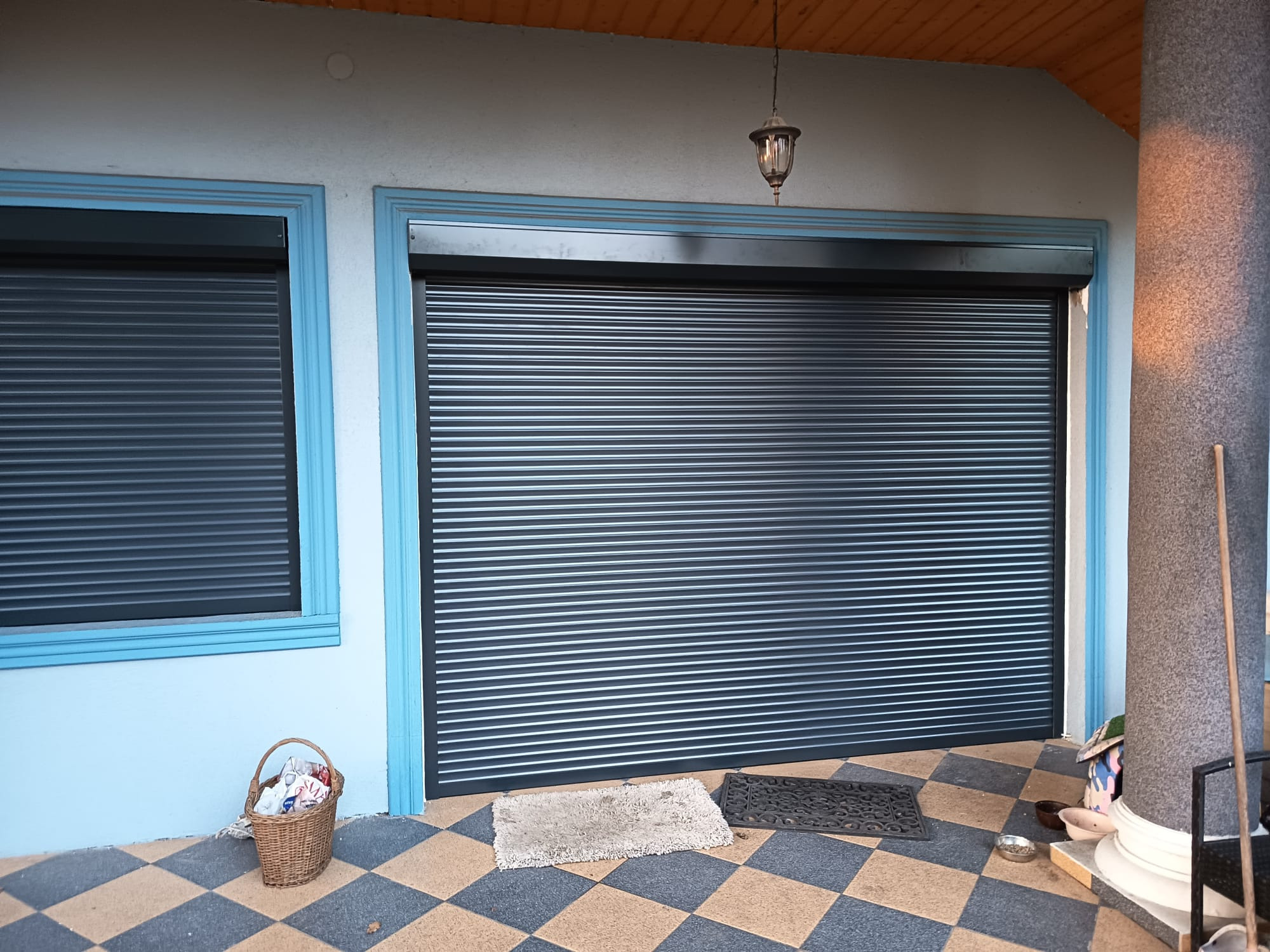 Zewnętrzna roleta antywłamaniowa w kolorze grafitowym, zamontowana na tle błękitnej ściany z oknem. Na podłodze dywaniki i koszyk.