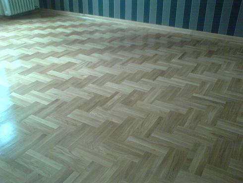 Jasna, parkietowa podłoga ułożona w jodełkę, z widocznym fragmentem ściany w pionowe pasy. Widoczne odbicie światła na powierzchni podłogi.