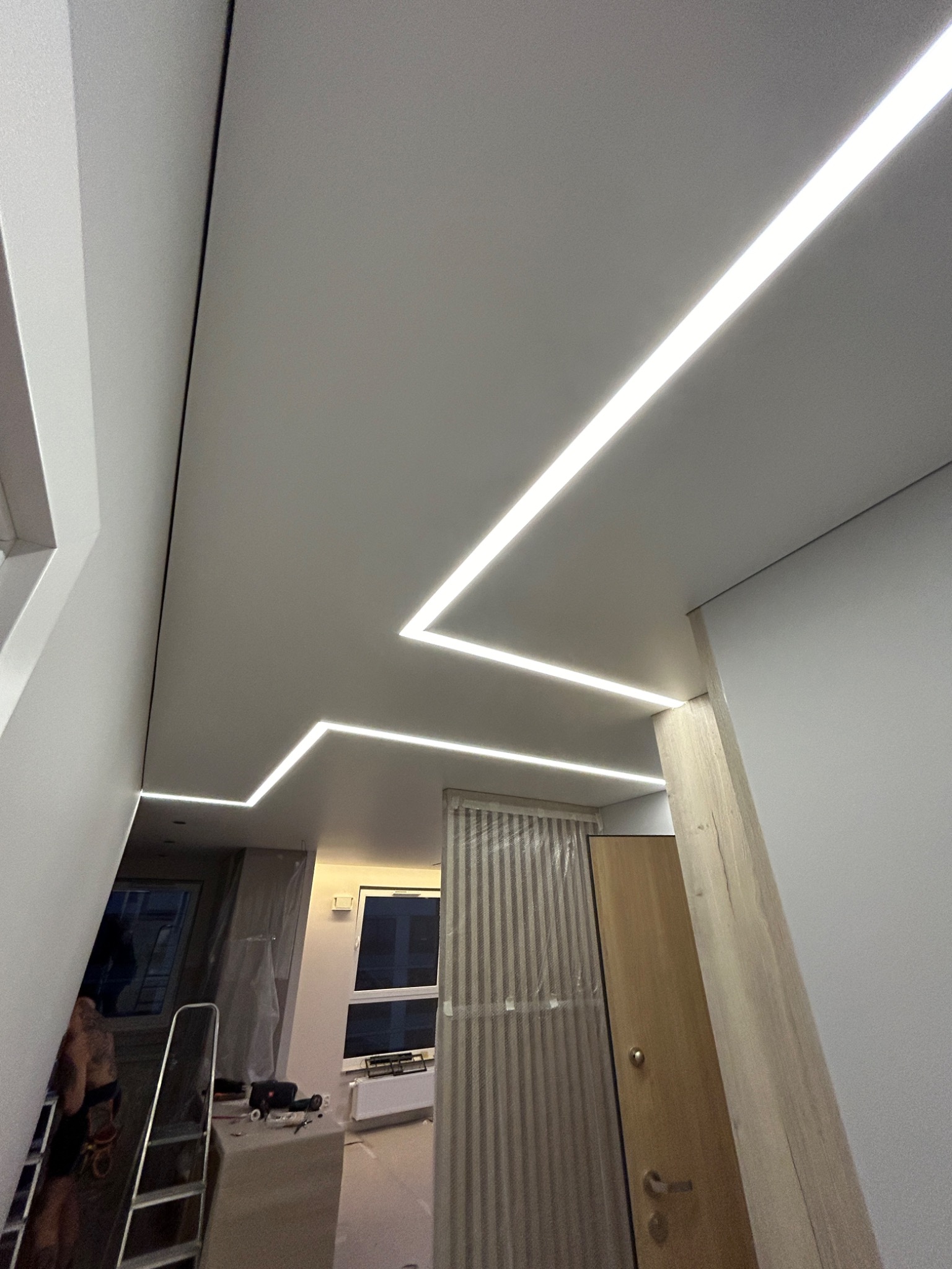 Biały sufit z wbudowanym, liniowym oświetleniem LED w kształcie zygzaka, widoczne elementy konstrukcyjne i fragment pomieszczenia w trakcie remontu z drabiną i folią ochronną.