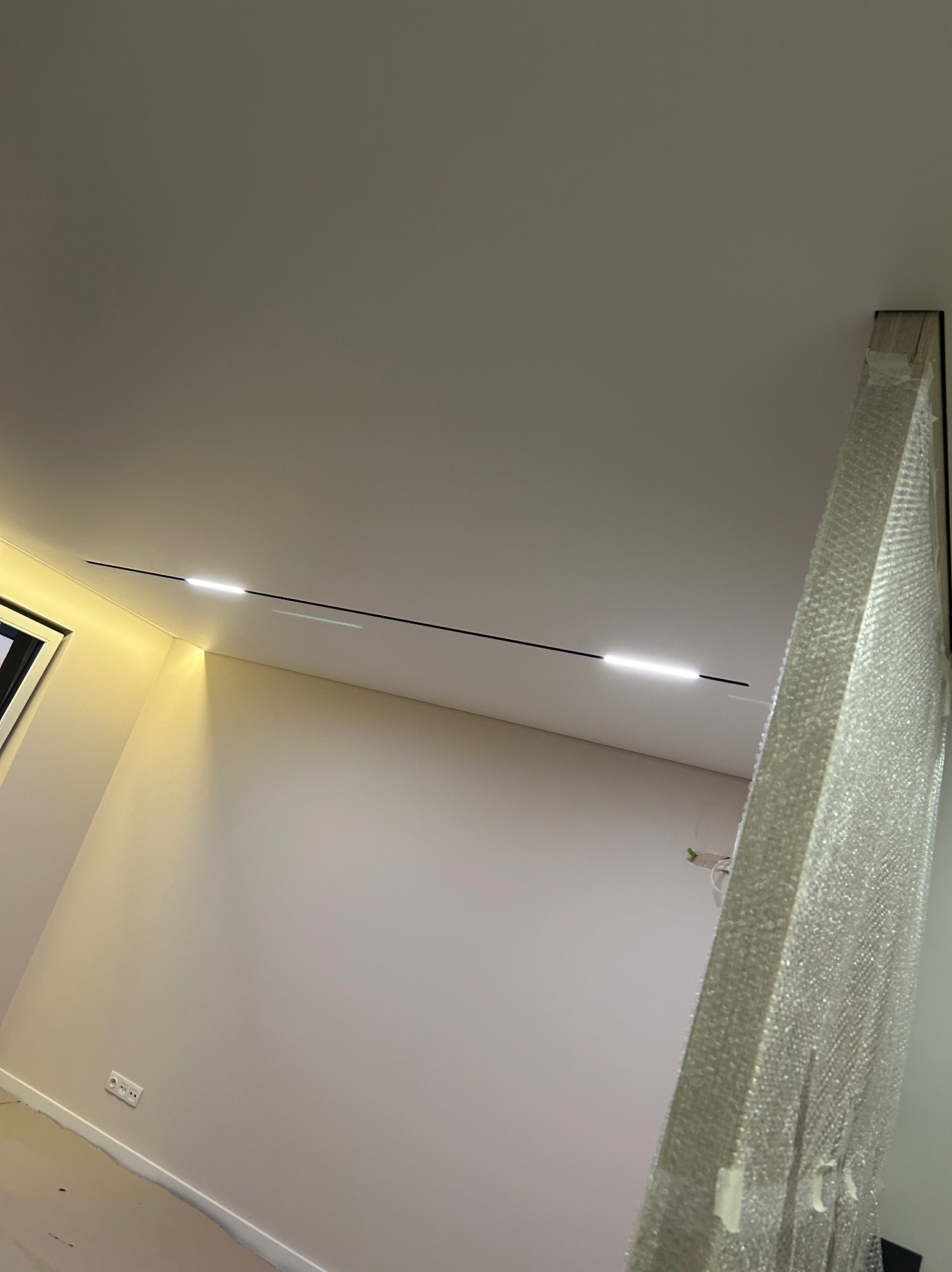 Nowoczesny sufit napinany z liniowym oświetleniem LED, widoczny fragment ściany z gniazdkiem elektrycznym i zabezpieczona folią budowlaną rama w rogu pomieszczenia.