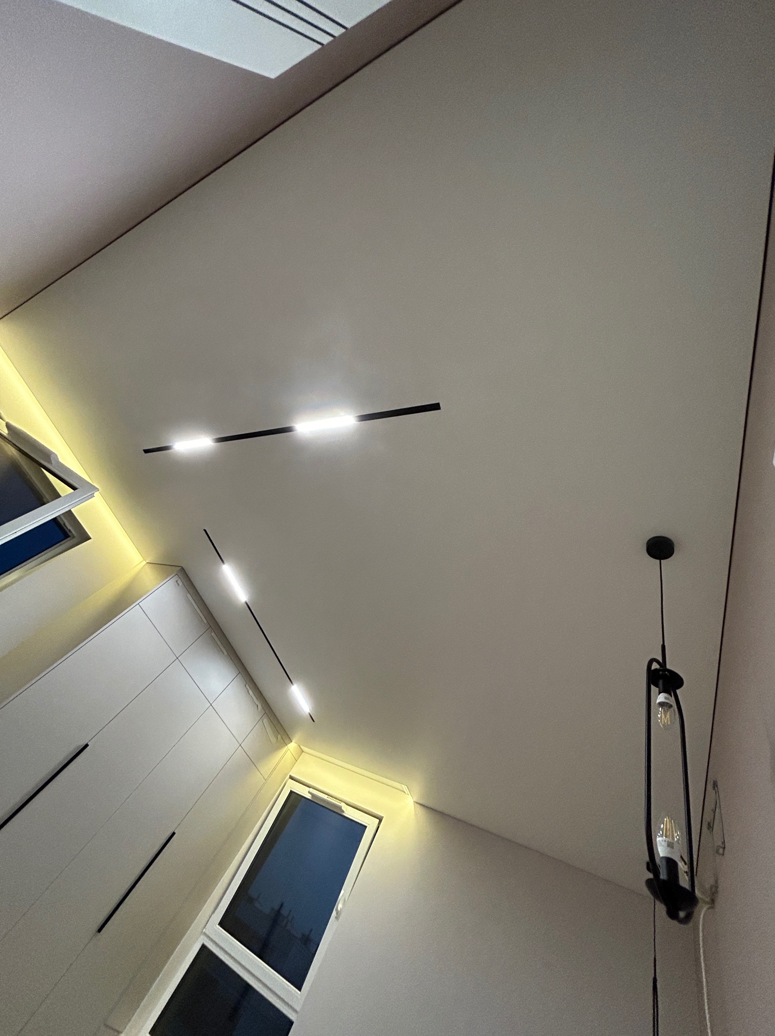 Jasny, gładki sufit z wbudowanym oświetleniem LED w postaci czarnych, prostych linii oraz wisząca, czarna lampa z odsłoniętymi żarówkami, widok z dołu.