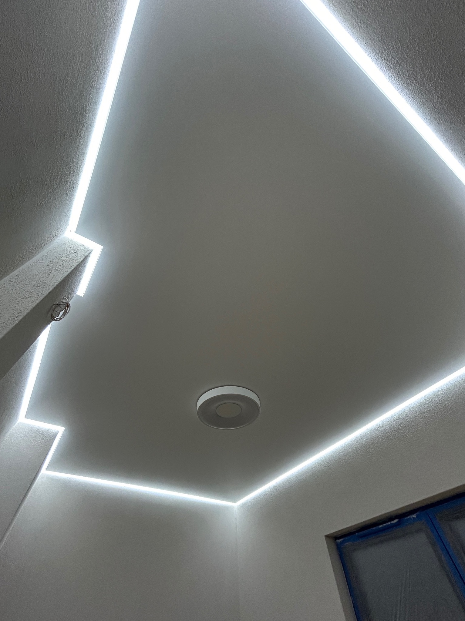 Biały sufit z oświetleniem LED w kształcie prostokąta, centralnie umieszczona okrągła lampa. Widoczne fragmenty okna zabezpieczonego folią malarską.