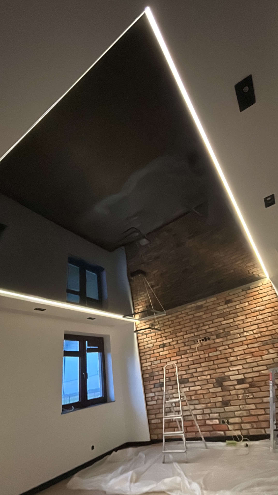 Czarny, błyszczący sufit z oświetleniem LED wzdłuż krawędzi, widoczny w trakcie montażu, z zabezpieczoną folią podłogą i drabiną opartą o ścianę z cegły.