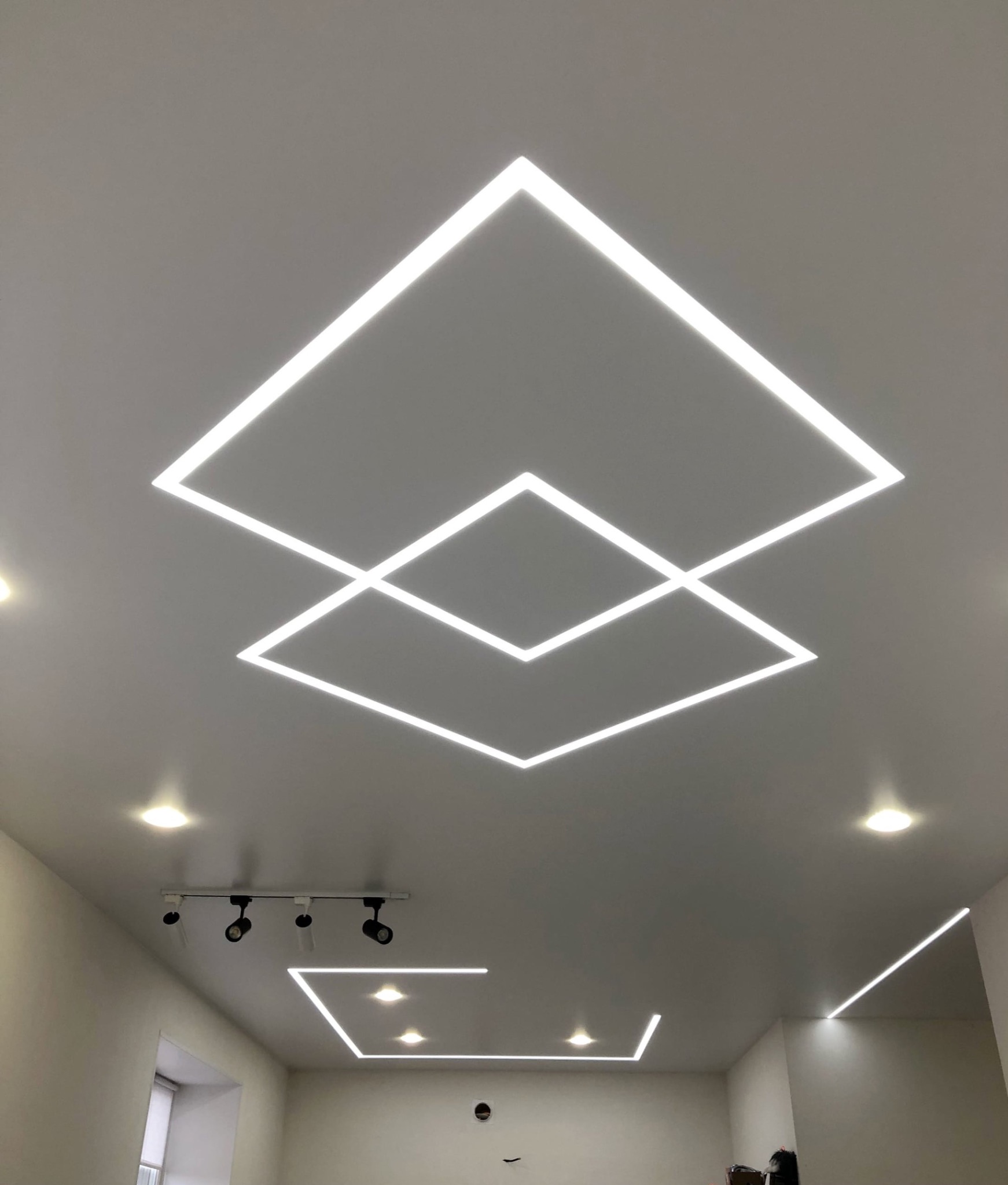 Biały sufit z geometrycznym wzorem z oświetlenia LED w kształcie dwóch nakładających się kwadratów, dodatkowe oświetlenie punktowe i szynowe.