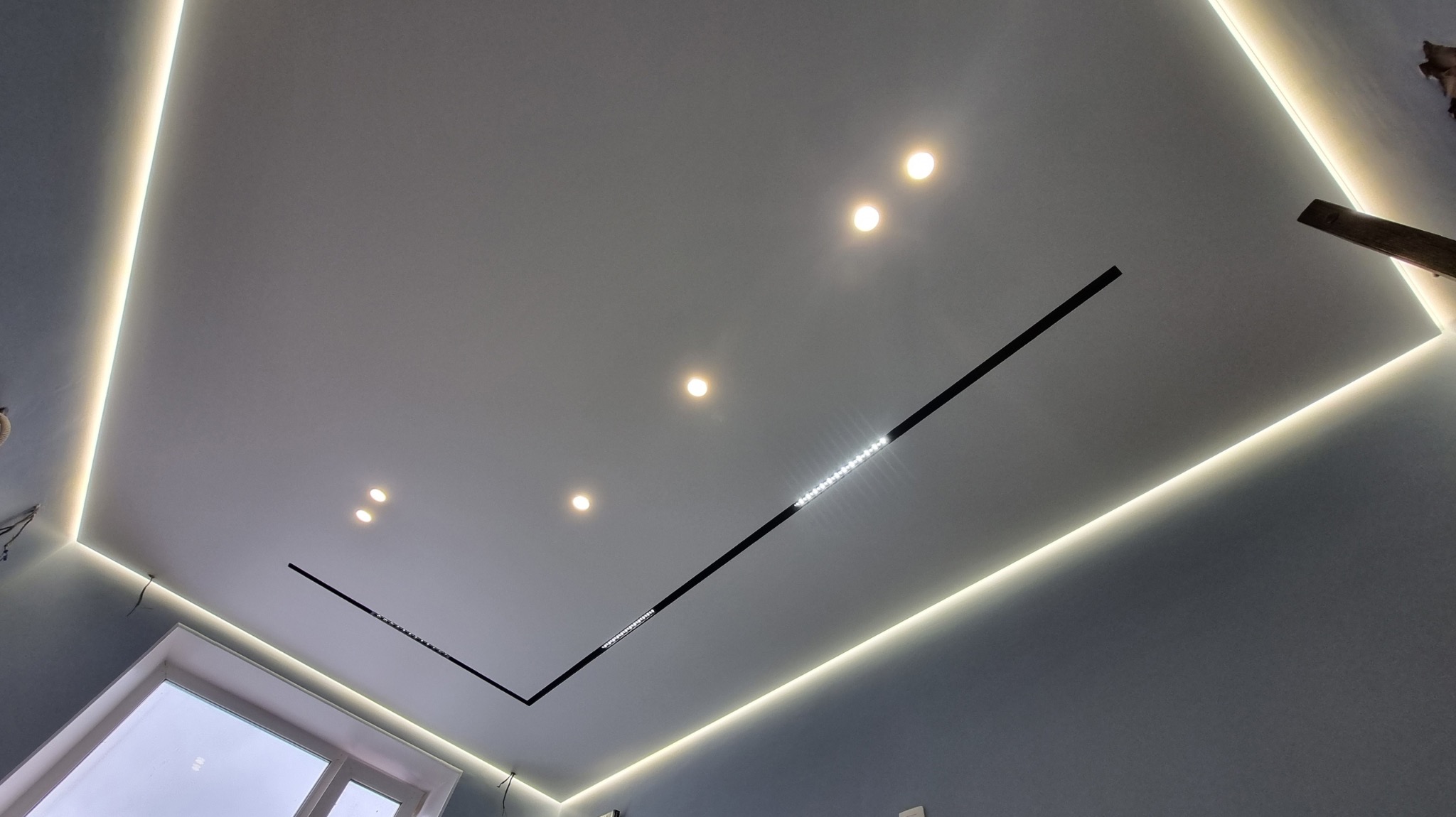 Nowoczesny sufit z oświetleniem LED obwodowym i liniowymi lampami LED, widoczne okno i fragment ściany.