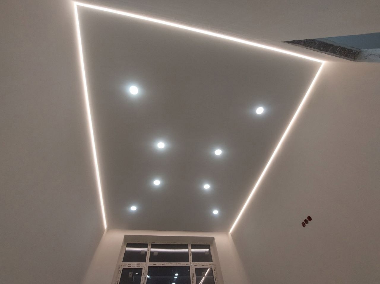 Biały sufit napinany z oświetleniem LED w kształcie trapezu i punktowymi lampami, widoczne okno w dolnej części kadru.