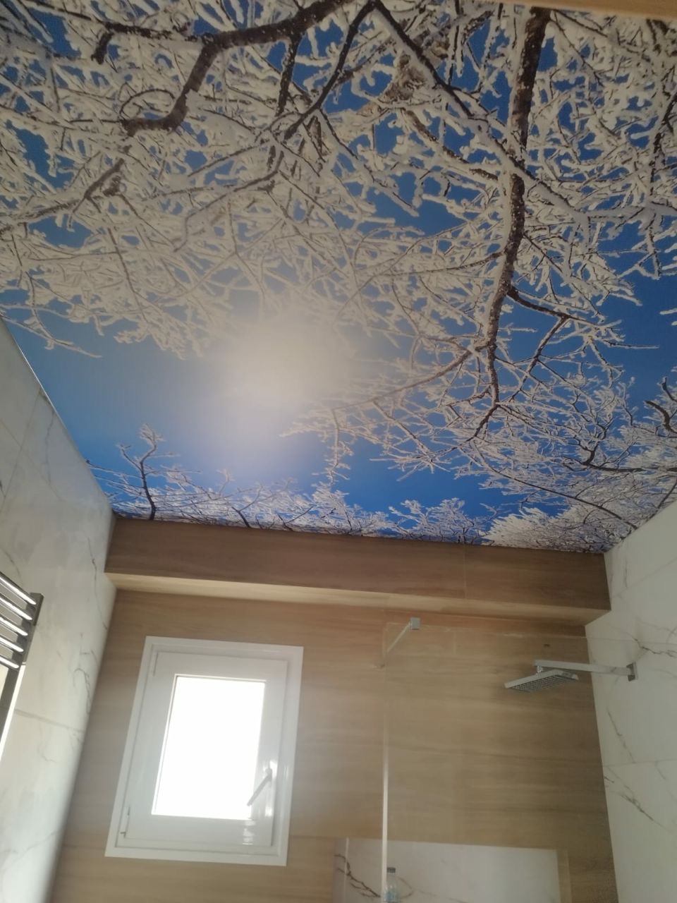 Łazienka z napinanym sufitem z motywem zimowego lasu i błękitnego nieba, widoczny biały grzejnik i kabina prysznicowa.