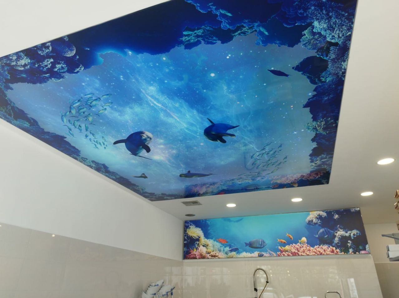 Niebieski sufit z nadrukiem 3D przedstawiającym podwodny świat z delfinami i rafą koralową, z dodatkowym panelem dekoracyjnym na ścianie w podobnym stylu.