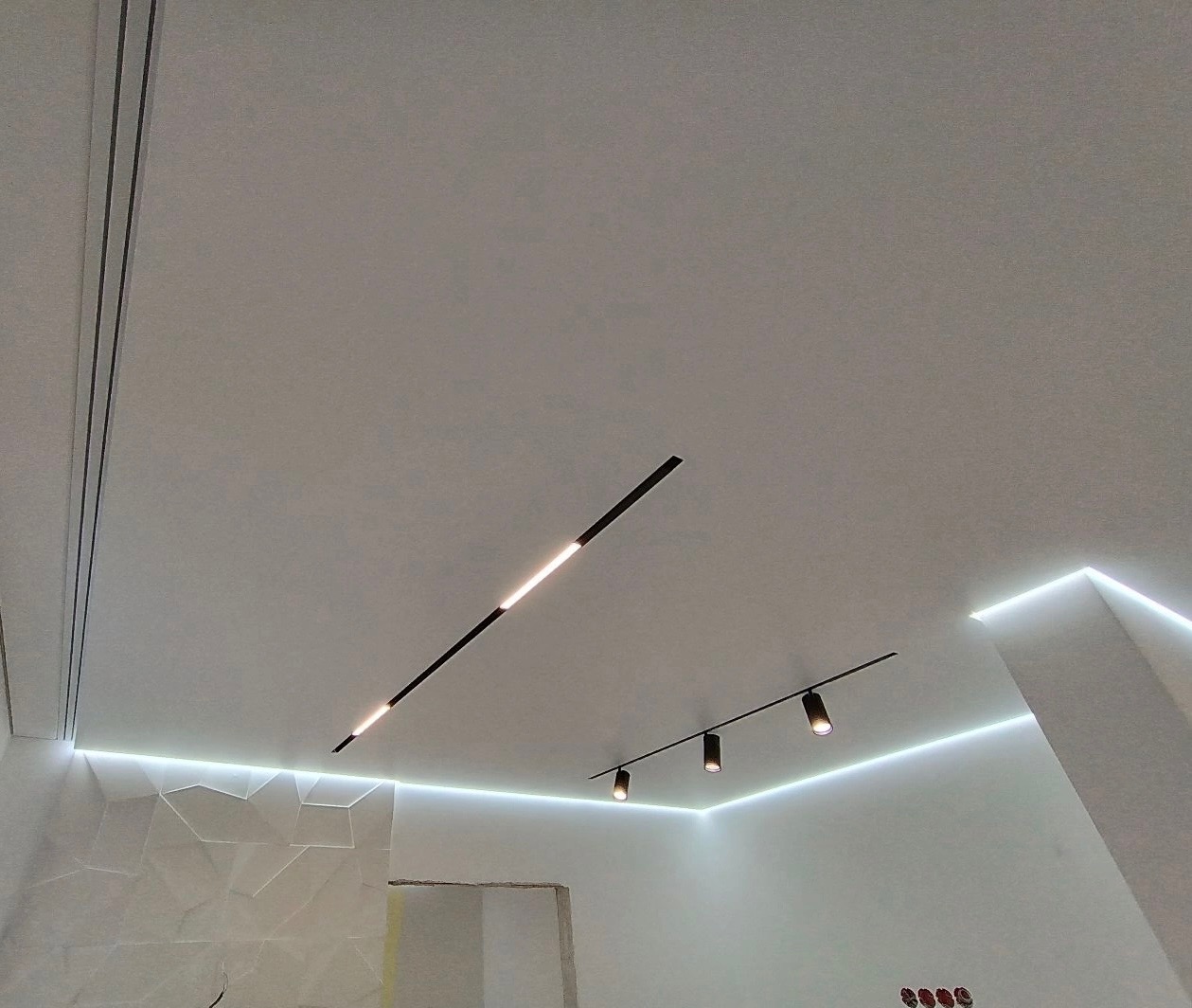 Biały sufit z oświetleniem LED: liniowym, punktowym na szynie i wzdłuż ściany. W tle widoczna ściana z dekoracyjną mozaiką.