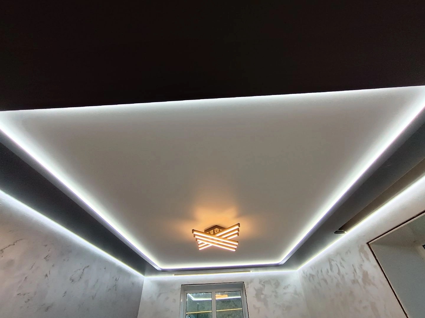 Biały, matowy sufit z podświetleniem LED wzdłuż krawędzi, centralnie umieszczona nowoczesna lampa sufitowa z geometrycznym wzorem, szare ściany z efektem dekoracyjnym.