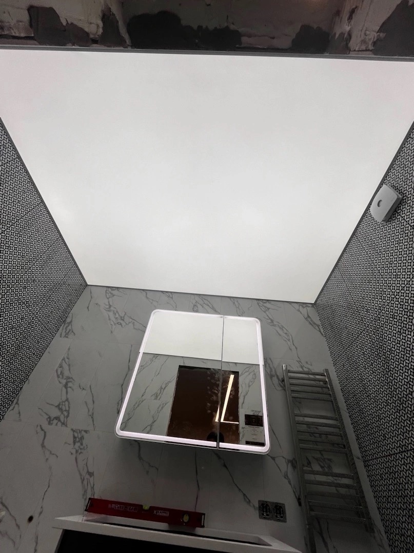 Biały, podświetlany sufit w łazience z płytkami imitującymi marmur i wzorzystymi, czarno-białymi płytkami, widoczny poziomica na umywalce i grzejnik drabinkowy. Widok z dołu, z lekkim nachyleniem.