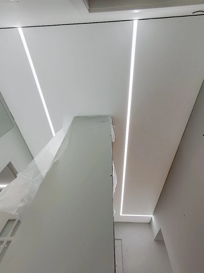 Biały sufit z liniowymi oprawami oświetleniowymi LED, widoczny fragment kolumny zabezpieczonej folią ochronną, w tle fragment okna.