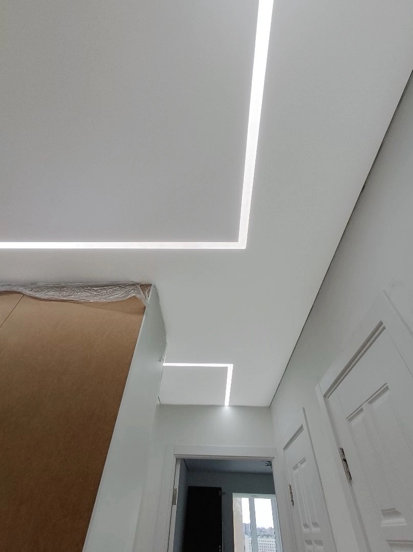 Biały sufit z wbudowanym oświetleniem LED w kształcie prostokątów, widok z dołu w korytarzu z białymi drzwiami i częściowo zasłoniętym meblem z folią ochronną.