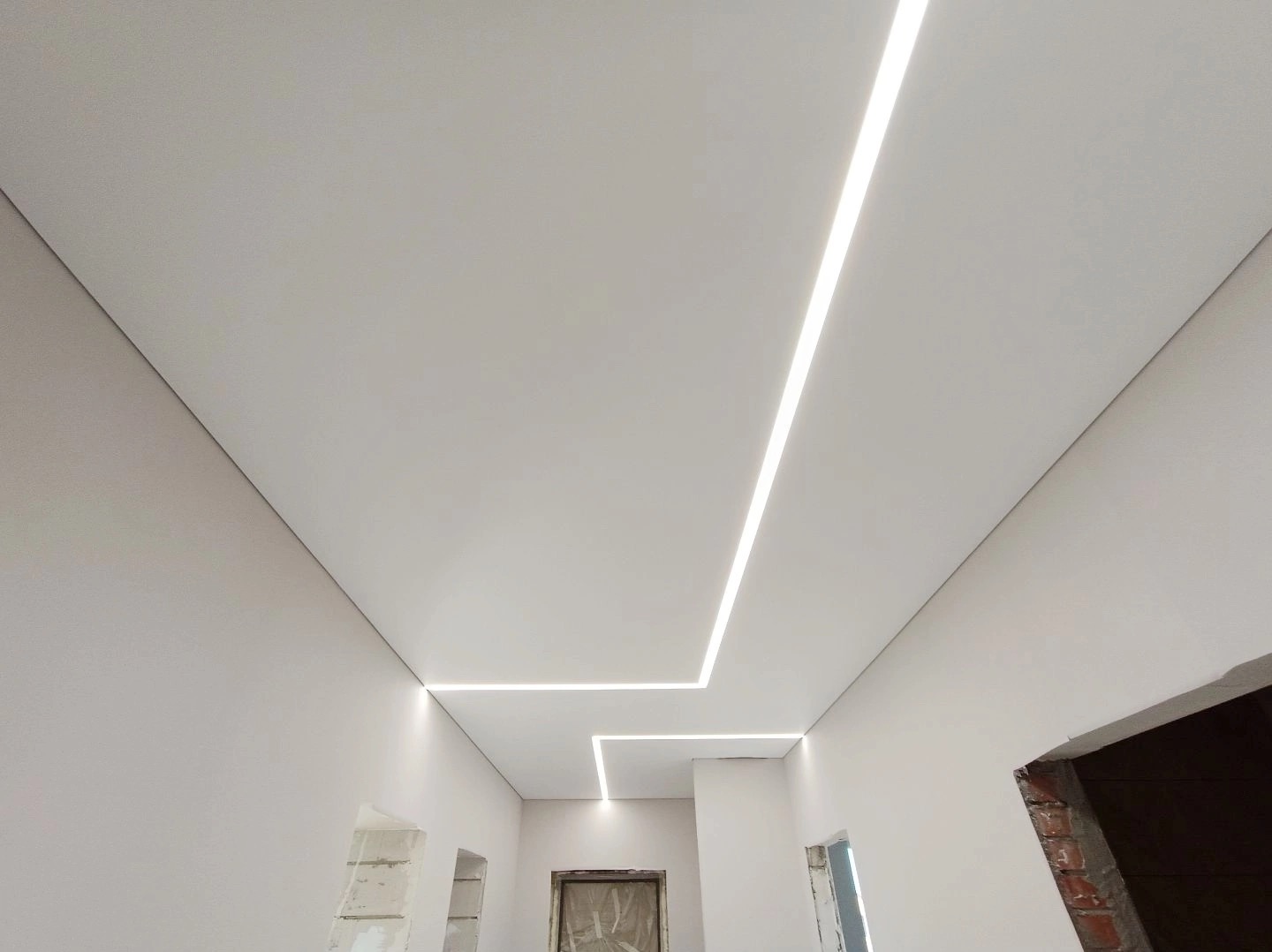 Biały sufit napinany z wbudowanym liniowym oświetleniem LED tworzącym geometryczny wzór, widoczne surowe ściany i otwory drzwiowe w trakcie remontu.