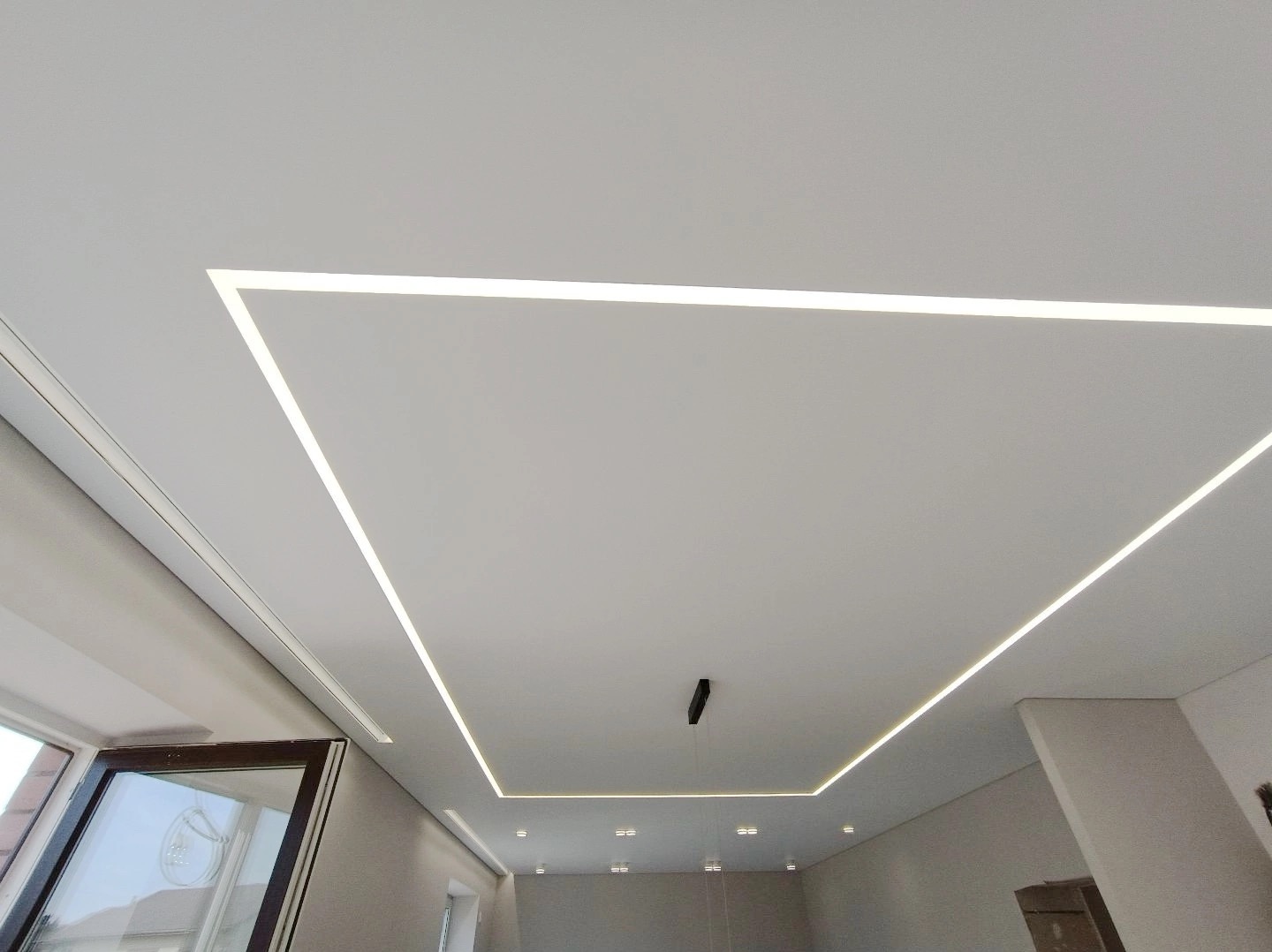 Biały sufit z napinanym obramowaniem z oświetleniem LED w kształcie kwadratu, widoczna lampa wisząca i fragment okna po lewej stronie.
