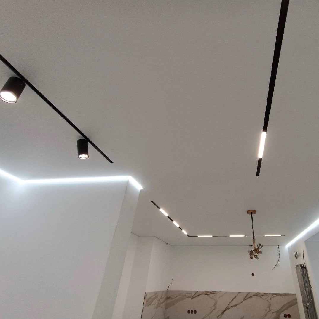 Biały sufit z różnymi rodzajami oświetlenia: reflektory na szynoprzewodach, listwy LED wzdłuż krawędzi oraz liniowe oprawy oświetleniowe. Widoczny fragment ściany z marmurowym wykończeniem.