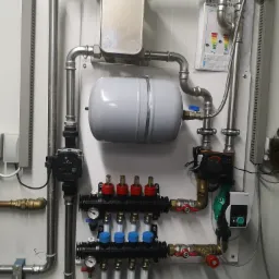 Rozdzielacz podłogowy z pompami obiegowymi, zbiornikiem wyrównawczym i wymiennikiem ciepła zamontowany na ścianie z blachy trapezowej, widoczne rury stalowe i miedziane, manometry i zawory.