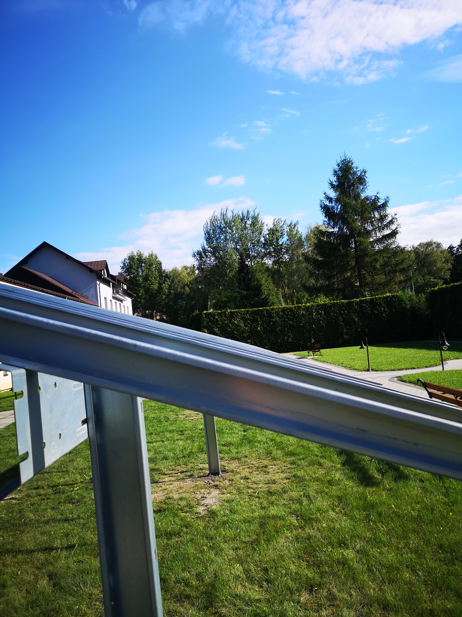 Metalowa konstrukcja nośna na trawniku, prawdopodobnie do montażu paneli słonecznych, z widokiem na budynek i zielony krajobraz w tle.