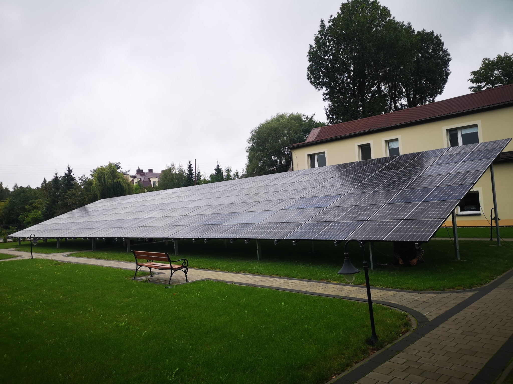 Instalacja paneli fotowoltaicznych w Goczałkowicach-Zdroju, widok na rozległą konstrukcję z paneli słonecznych zamontowaną obok budynku na trawniku, z ławką i lampami parkowymi w tle, pochmurne niebo.