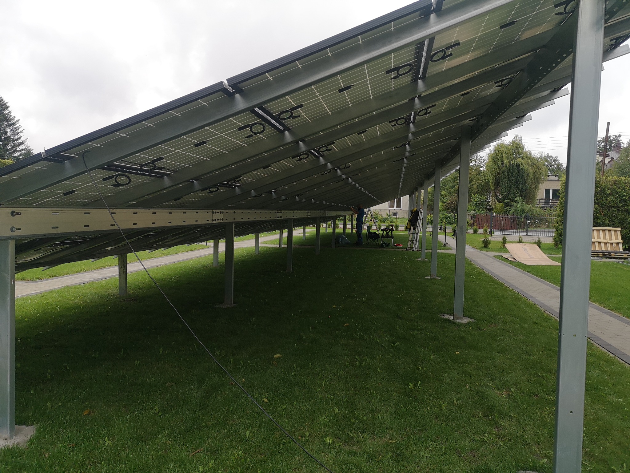 Widok od spodu na konstrukcję paneli fotowoltaicznych zamontowanych na metalowych słupach na trawniku, dwóch instalatorów pracuje w tle.