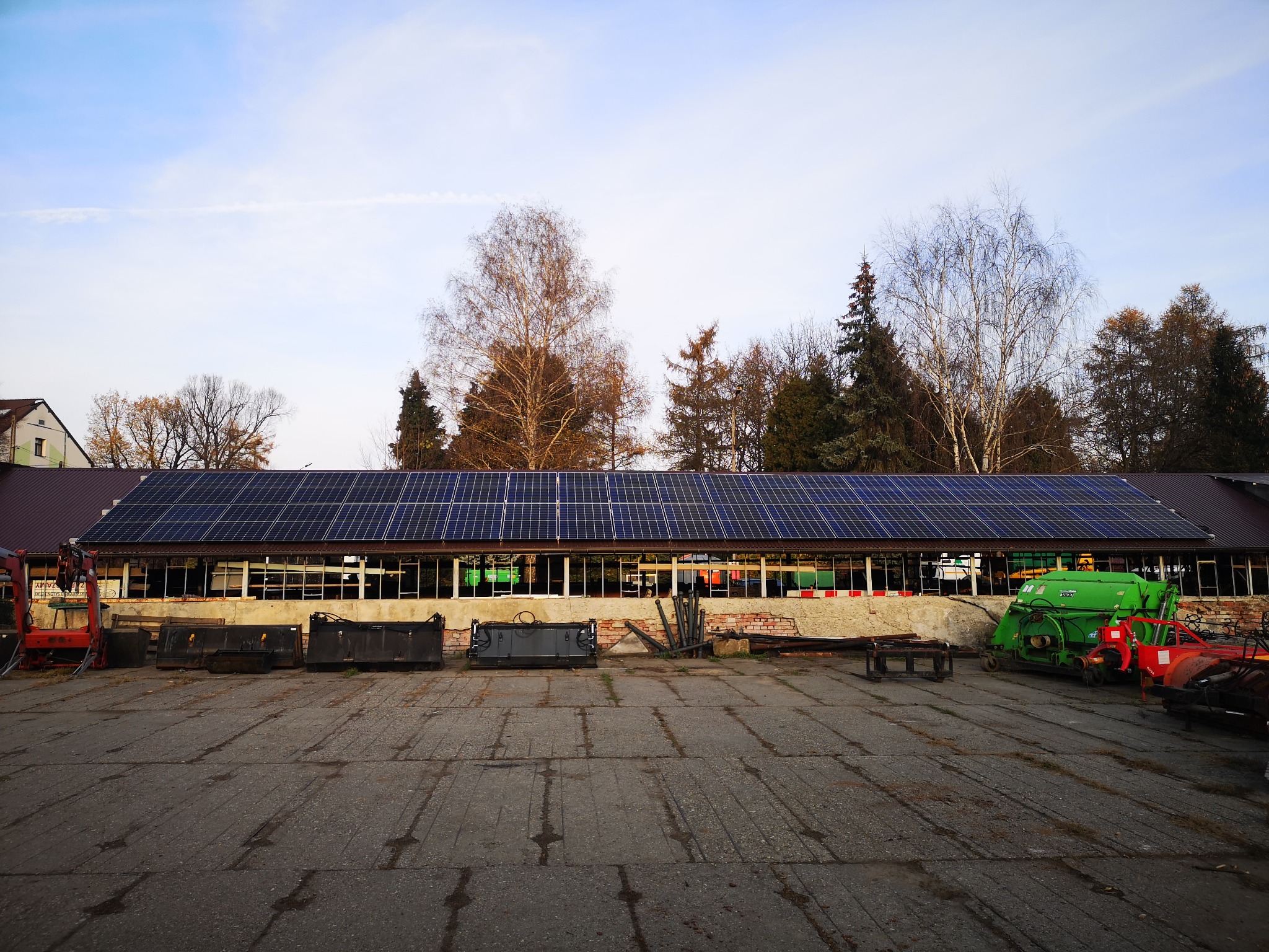 Budynek gospodarczy z panelami słonecznymi na dachu, widoczne maszyny rolnicze i sprzęt przed budynkiem.