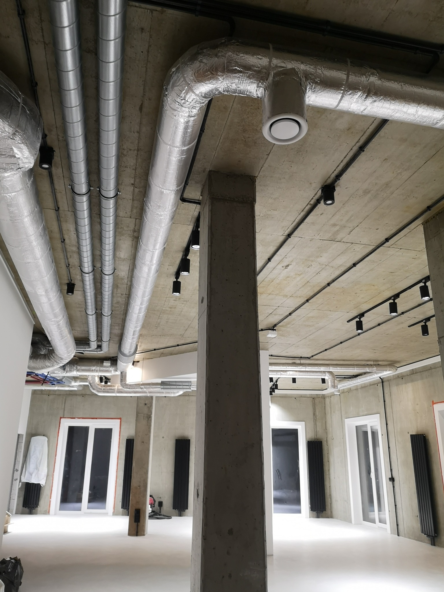 Surowy betonowy sufit z widocznymi przewodami wentylacyjnymi w srebrnej izolacji, czarnymi szynami oświetleniowymi i okrągłymi dyfuzorami w nowym biurze.