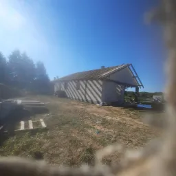 Budowa domu w stanie surowym otwartym, widoczne ściany z bloczków, więźba dachowa, palety i materiały budowlane na działce, nieostre ujęcie przez przeszkodę.