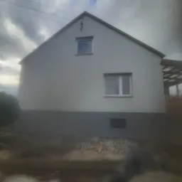 Boczny widok domu z jasną elewacją i ciemnoszarą podmurówką w pochmurny dzień, widoczny fragment drewnianej pergoli z boku.