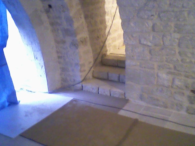 Kamienne schody w łukowatym przejściu, zabezpieczone kartonem na podłodze. Surowe, jasne kamienne ściany i stopnie. W tle widoczne światło dzienne.