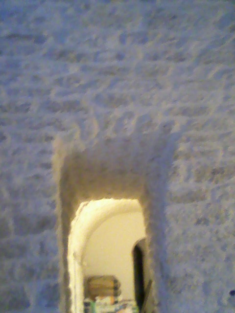 Niedokończone przejście w ścianie z surowego muru, widoczne wnętrze z meblami i przedmiotami. Oświetlenie wewnątrz pomieszczenia.