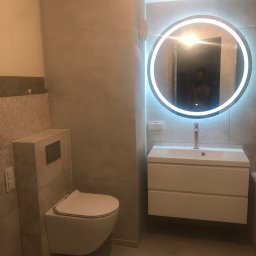 Serz-Bud - Wykończona łazienka z wiszącą toaletą, umywalką z szafką i okrągłym lustrem LED, widoczne szare płytki imitujące beton na ścianach i podłodze, odbicie fotografa w lustrze.