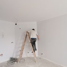 Serz-Bud - Malarz na drewnianej drabinie, w trakcie pracy w narożniku pomalowanego na biało pokoju, widoczne kable elektryczne wystające ze ściany, narzędzia malarskie i plamy farby na podłodze.