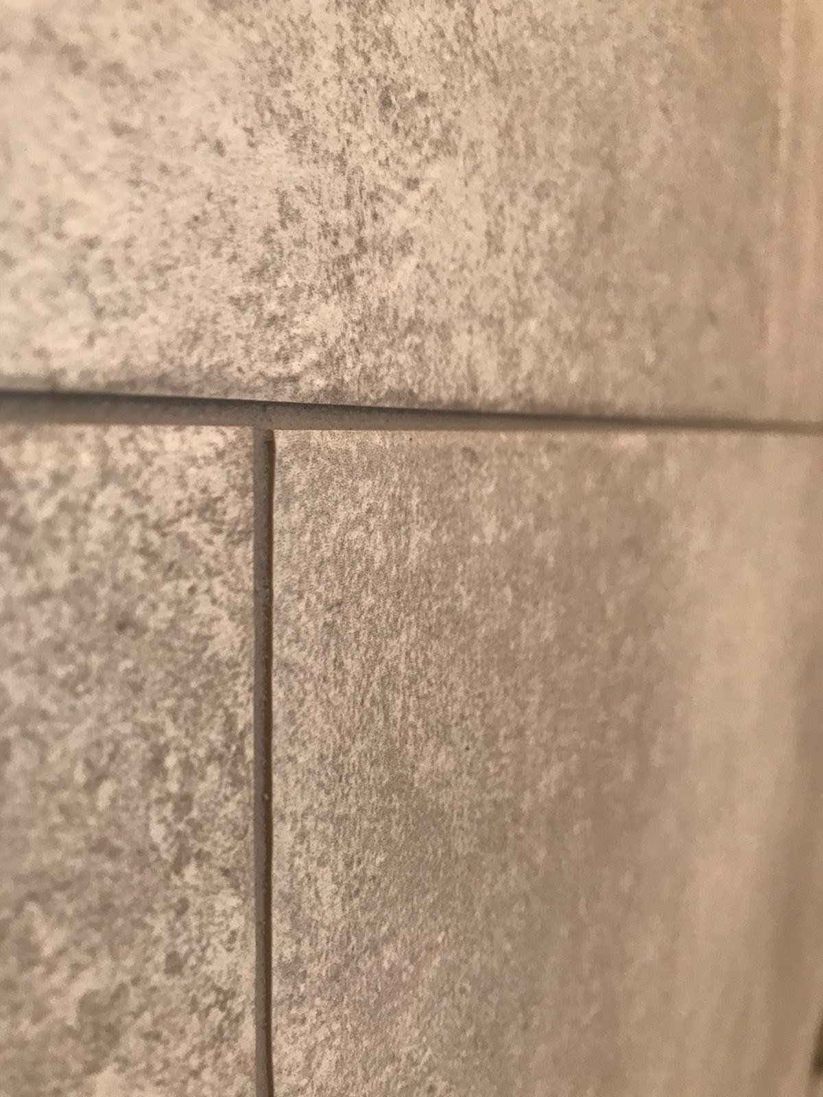 Zbliżenie na szare, matowe płytki ceramiczne na ścianie, z widocznymi fugami w odcieniu zbliżonym do płytek, ukazujące teksturę i wykończenie powierzchni.