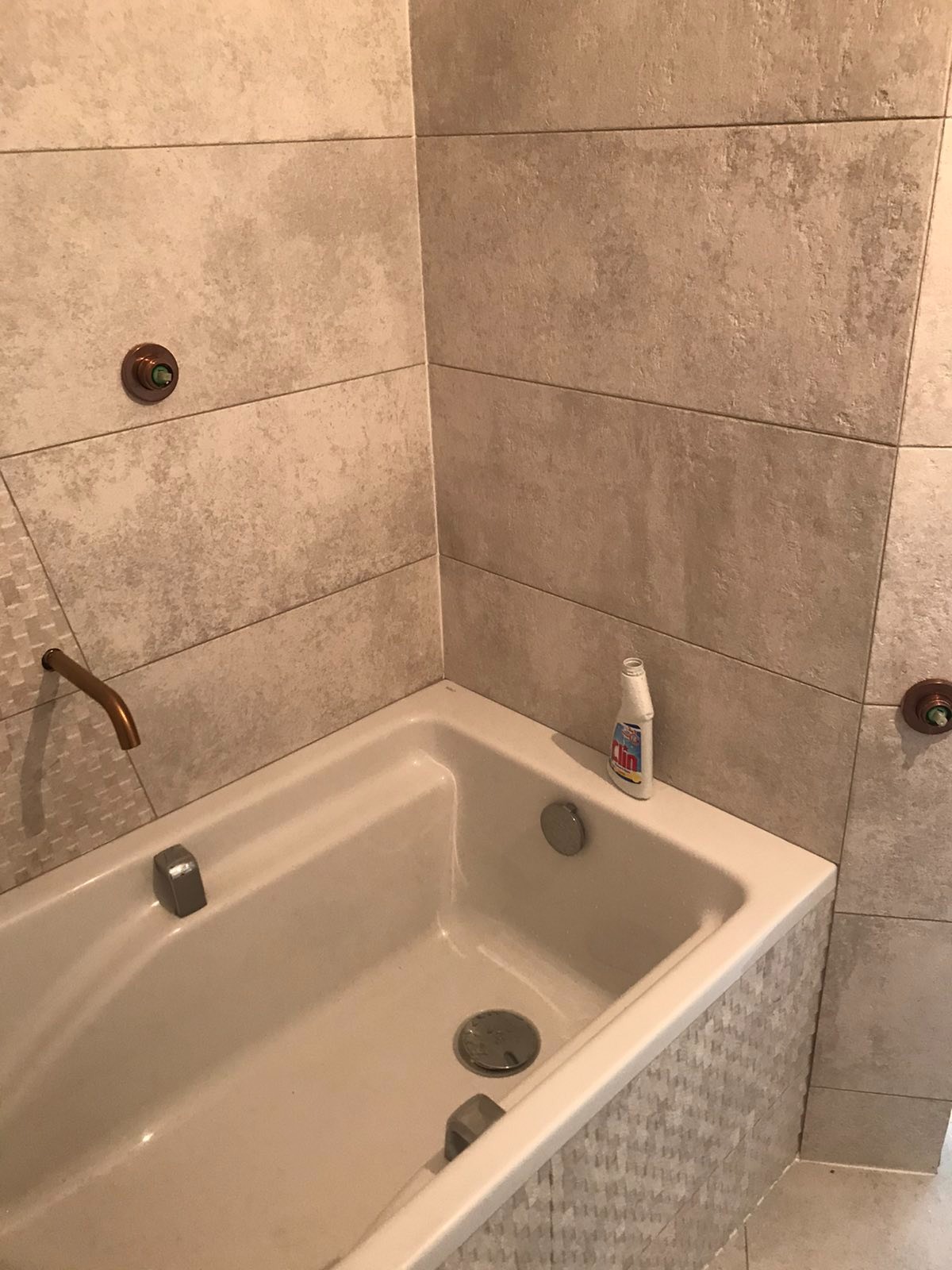 Beżowa wanna z baterią w kolorze miedzi, otoczona płytkami w odcieniach beżu i szarości, z butelką środka czyszczącego na krawędzi.