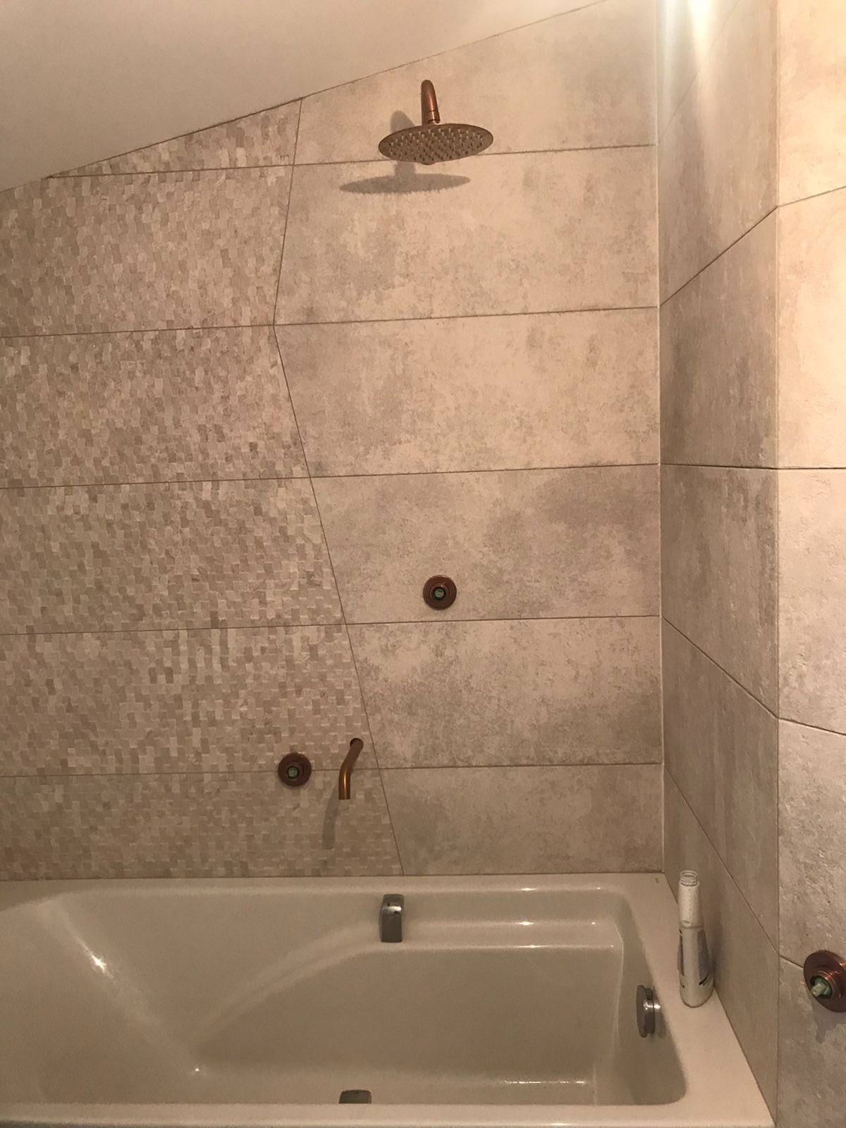 Wykończona łazienka z wanną, beżowymi płytkami o zróżnicowanej fakturze i miedzianą armaturą, w tym deszczownicą i kurkami, oświetlona górnym światłem.