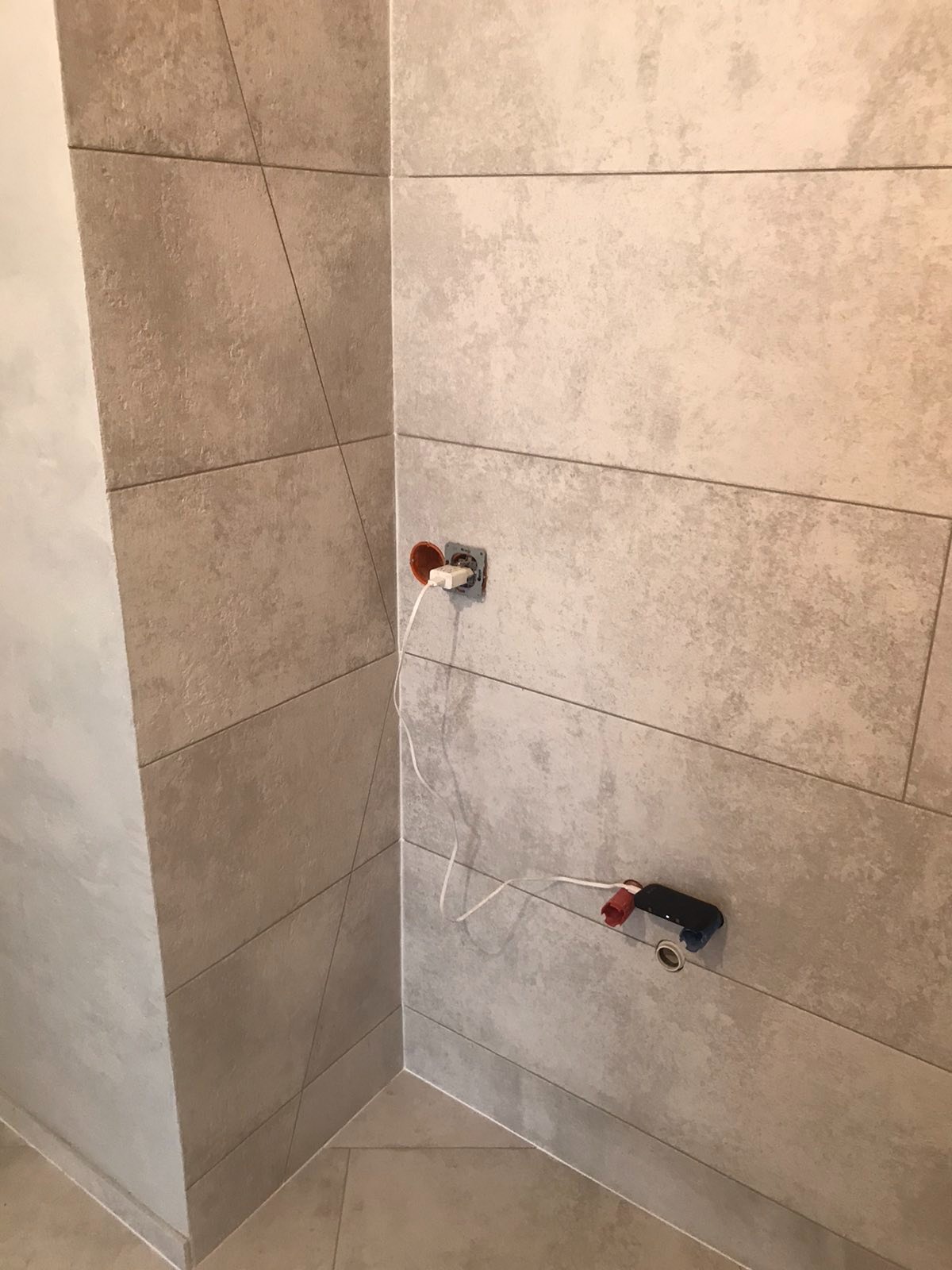 Ściana łazienki wyłożona beżowymi, podłużnymi płytkami ceramicznymi. Widoczne niedokończone podłączenia elektryczne i hydrauliczne, wystające przewody. Ułożenie płytek w rogu pomieszczenia...