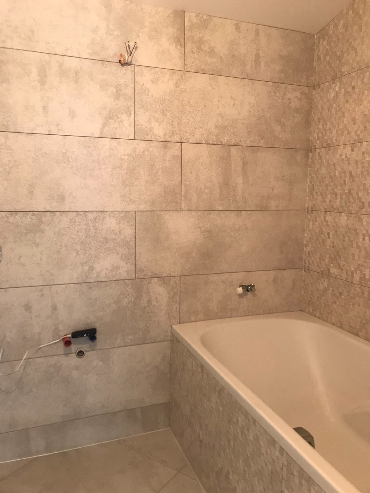 Ściana łazienki wyłożona beżowymi, prostokątnymi płytkami imitującymi kamień, z widoczną instalacją wodną i częścią wanny z mozaikową obudową.