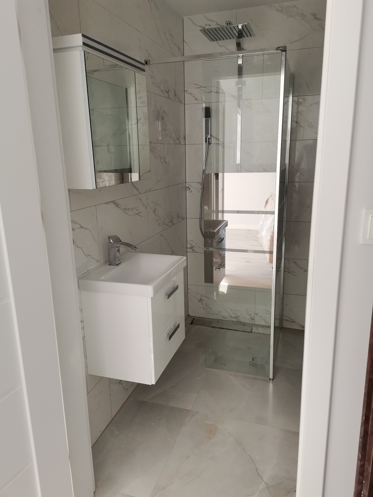 Nowoczesna łazienka po remoncie: biała umywalka z szafką, lustro w zabudowie, przeszklona kabina prysznicowa typu walk-in z deszczownicą, marmurowe płytki na ścianach i podłodze.