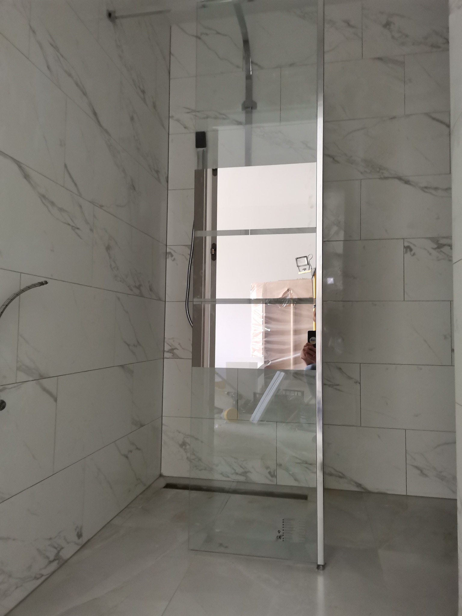 Szklana kabina prysznicowa typu walk-in z chromowanymi profilami, zamontowana w łazience wykończonej białymi płytkami imitującymi marmur, widoczny odpływ liniowy w podłodze.