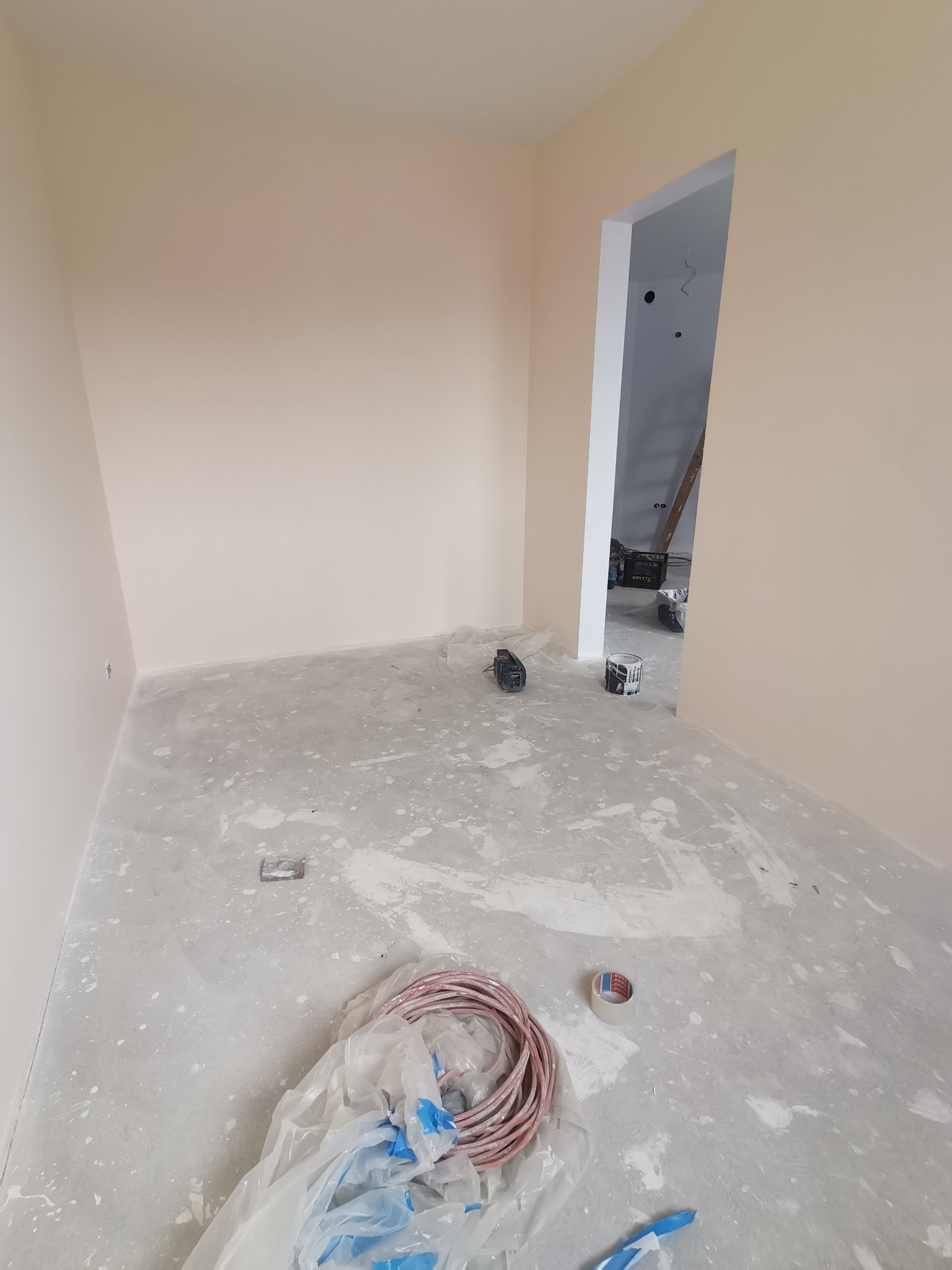 Świeżo pomalowane na beżowo ściany w pustym pokoju w trakcie remontu, widoczne ślady farby na podłodze, zwój kabla w folii, puszka z farbą i narzędzia w tle.