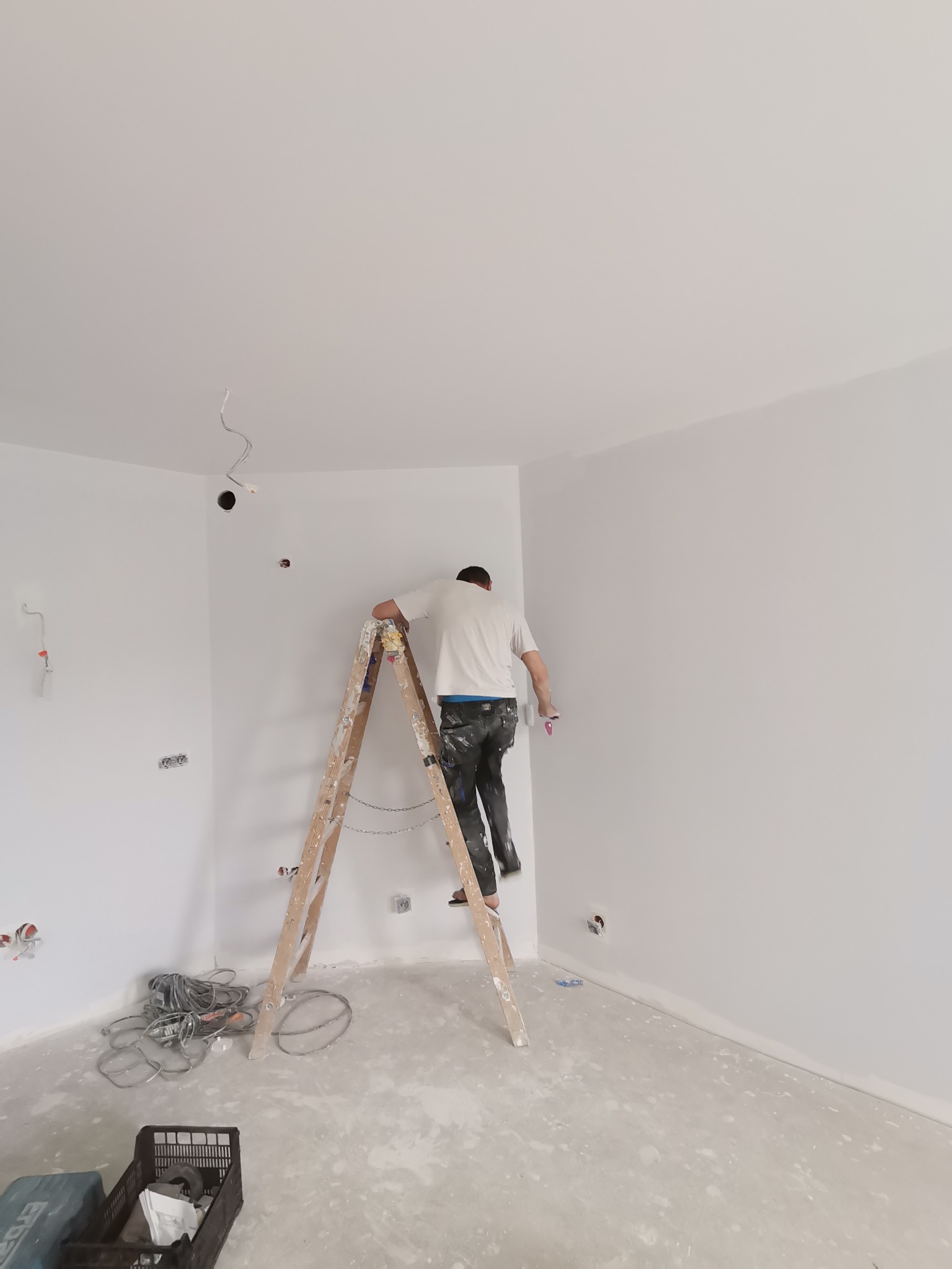 Malarz na drewnianej drabinie, w trakcie pracy w narożniku pomalowanego na biało pokoju, widoczne kable elektryczne wystające ze ściany, narzędzia malarskie i plamy farby na podłodze.