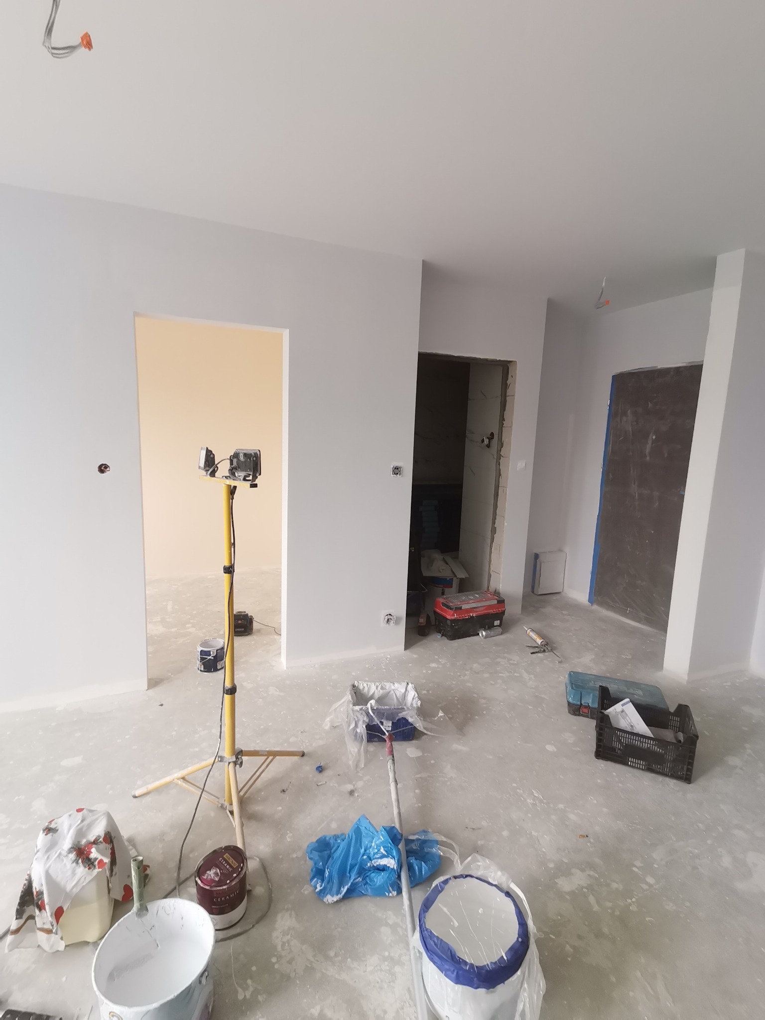 Wnętrze w trakcie odświeżania, z widocznymi śladami farby na podłodze, narzędziami malarskimi, lampą budowlaną i świeżo pomalowanymi ścianami na szaro.