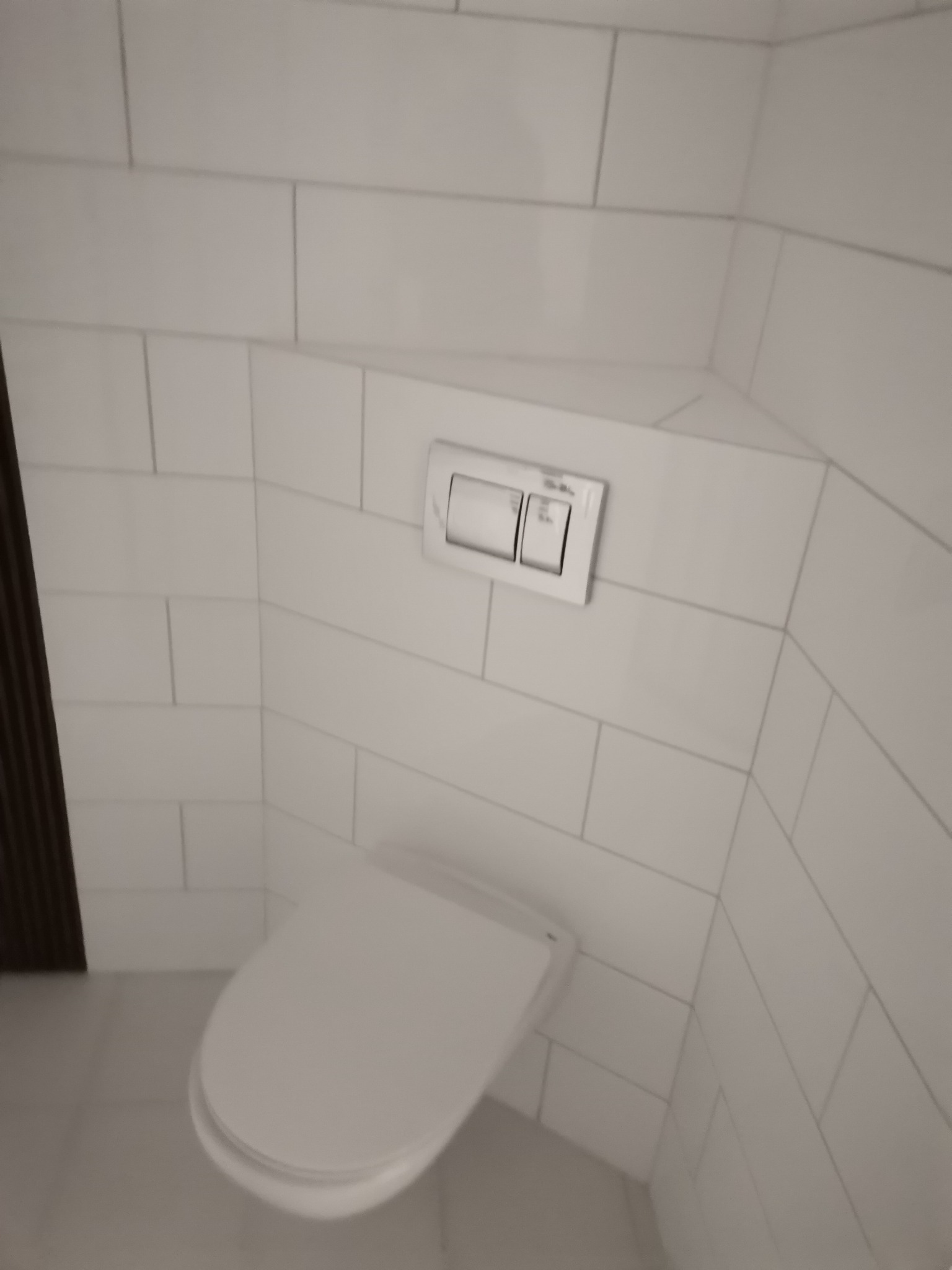 Nowoczesna toaleta wisząca z białymi płytkami ceramicznymi ułożonymi w regularny wzór, widoczny przycisk spłukiwania na ścianie, minimalistyczny design.