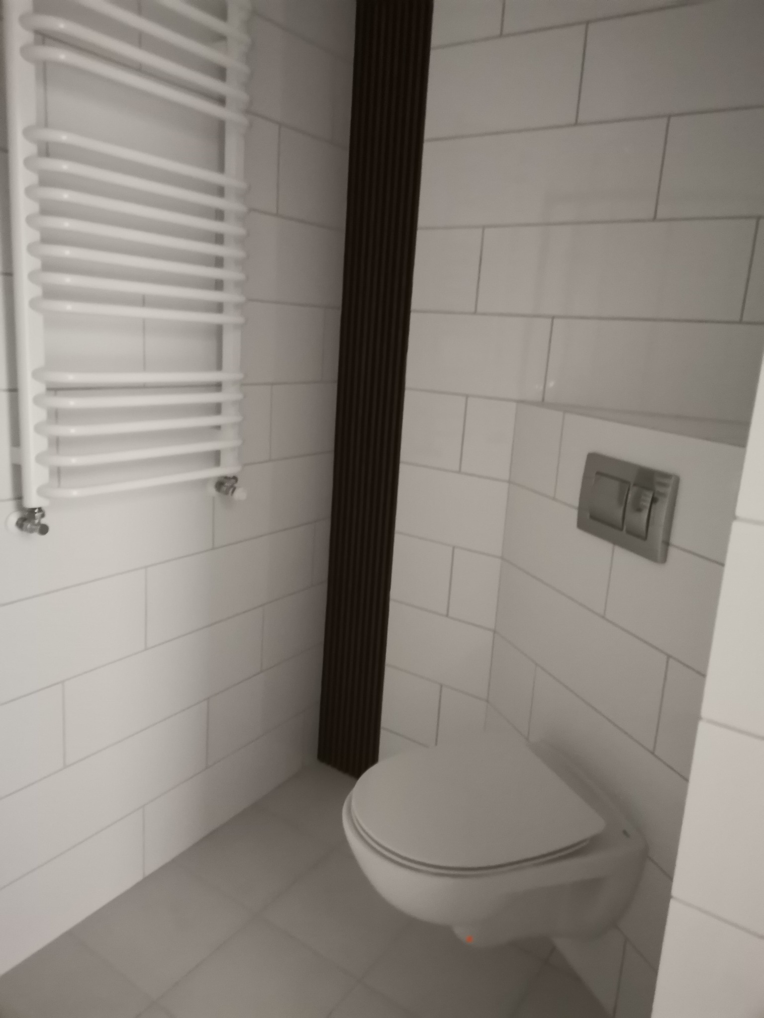 Nowoczesna łazienka z białymi, prostokątnymi płytkami, wiszącą toaletą z chromowanym przyciskiem spłukiwania, grzejnikiem drabinkowym oraz dekoracyjnym, ciemnobrązowym panelem ściennym.