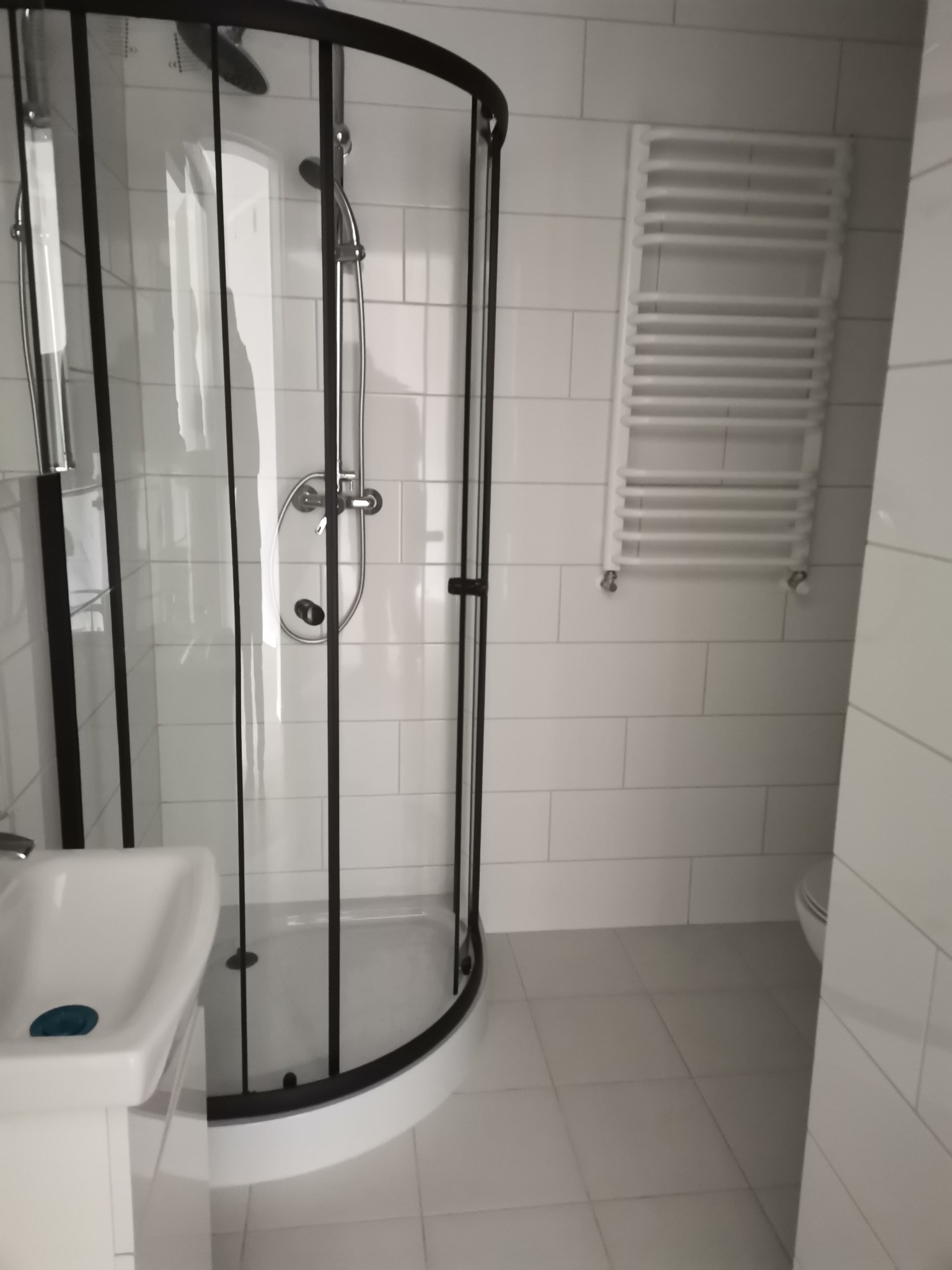 Wykończona łazienka w bieli z czarną kabiną prysznicową typu walk-in, grzejnik drabinkowy i umywalka z niebieskim akcentem.