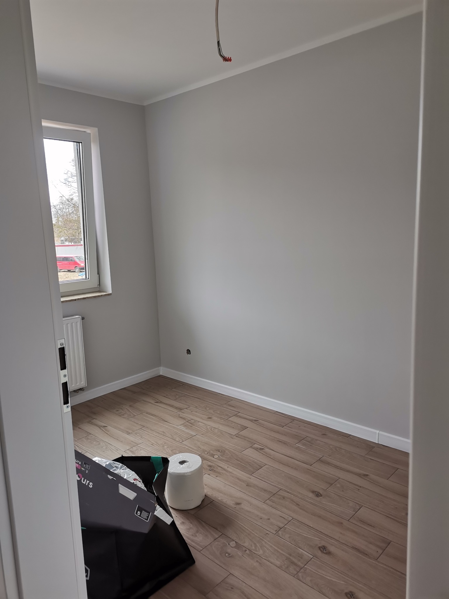 Pokój po malowaniu na szaro, z drewnopodobną podłogą, widoczne ślady po remoncie: karton, rolka papieru malarskiego.