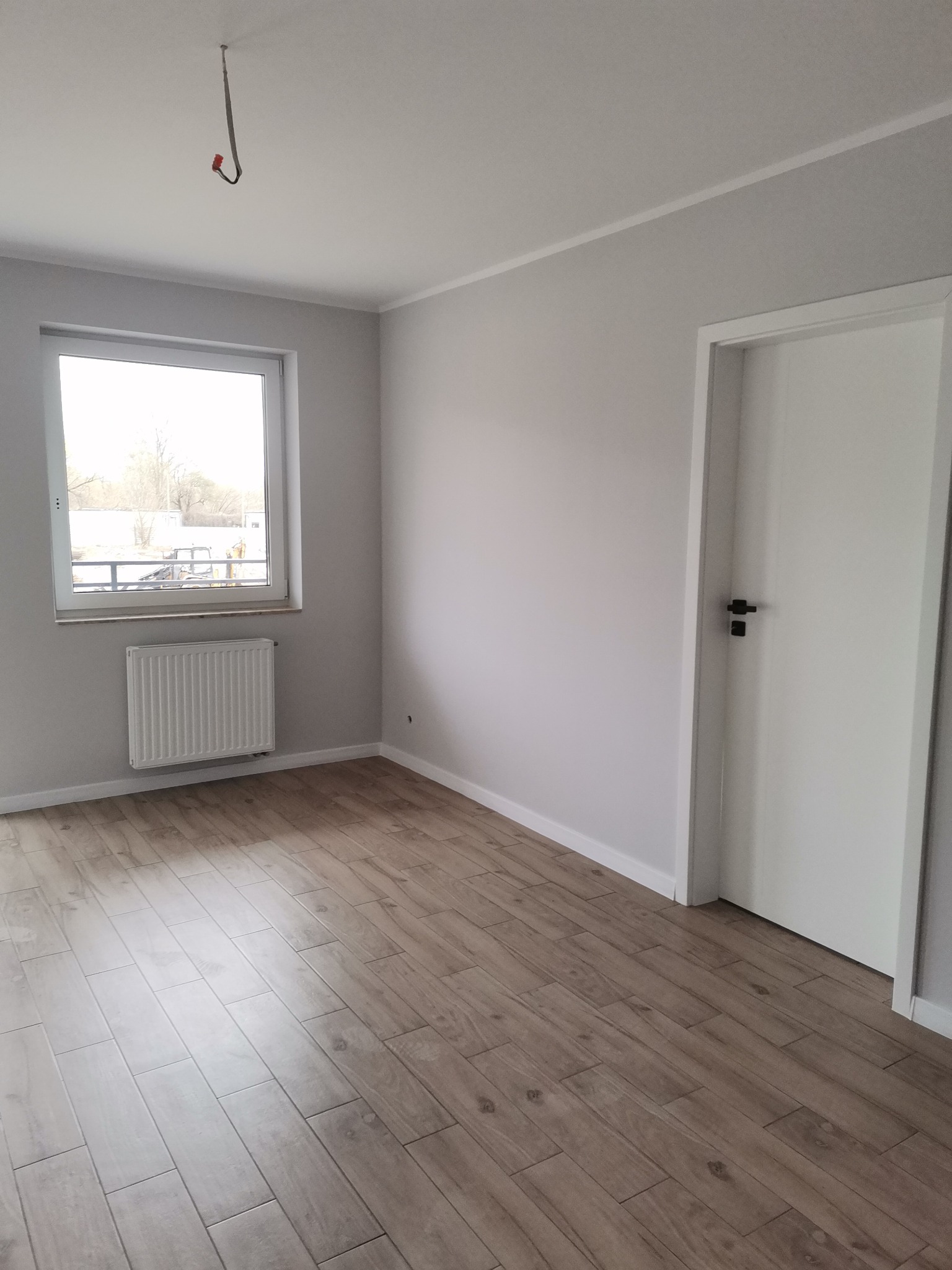 Pusty, jasny pokój po remoncie z panelami podłogowymi imitującymi drewno, białymi ścianami, grzejnikiem pod oknem i białymi drzwiami z czarną klamką.