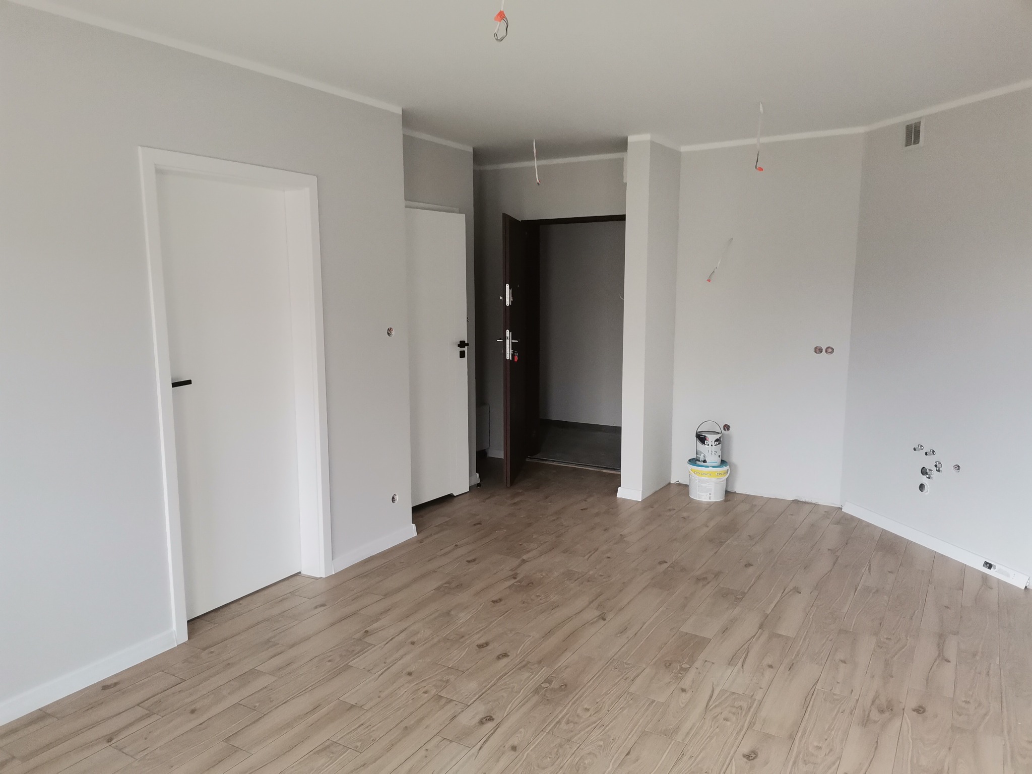 Wnętrze po odświeżeniu w Warszawie: świeżo pomalowane szare ściany, drewnopodobne panele podłogowe, białe drzwi i listwy przypodłogowe, widoczne puszki z farbą w rogu pomieszczenia.