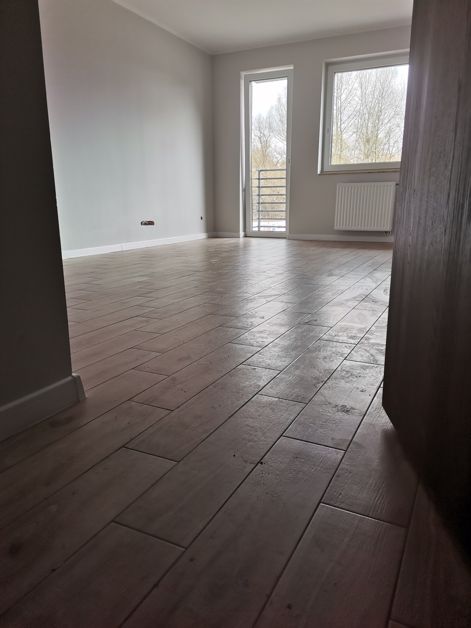 Ułożona podłoga z płytek imitujących drewno w pustym, jasnym pokoju z balkonem i grzejnikiem pod oknem, widok zza framugi drzwi.