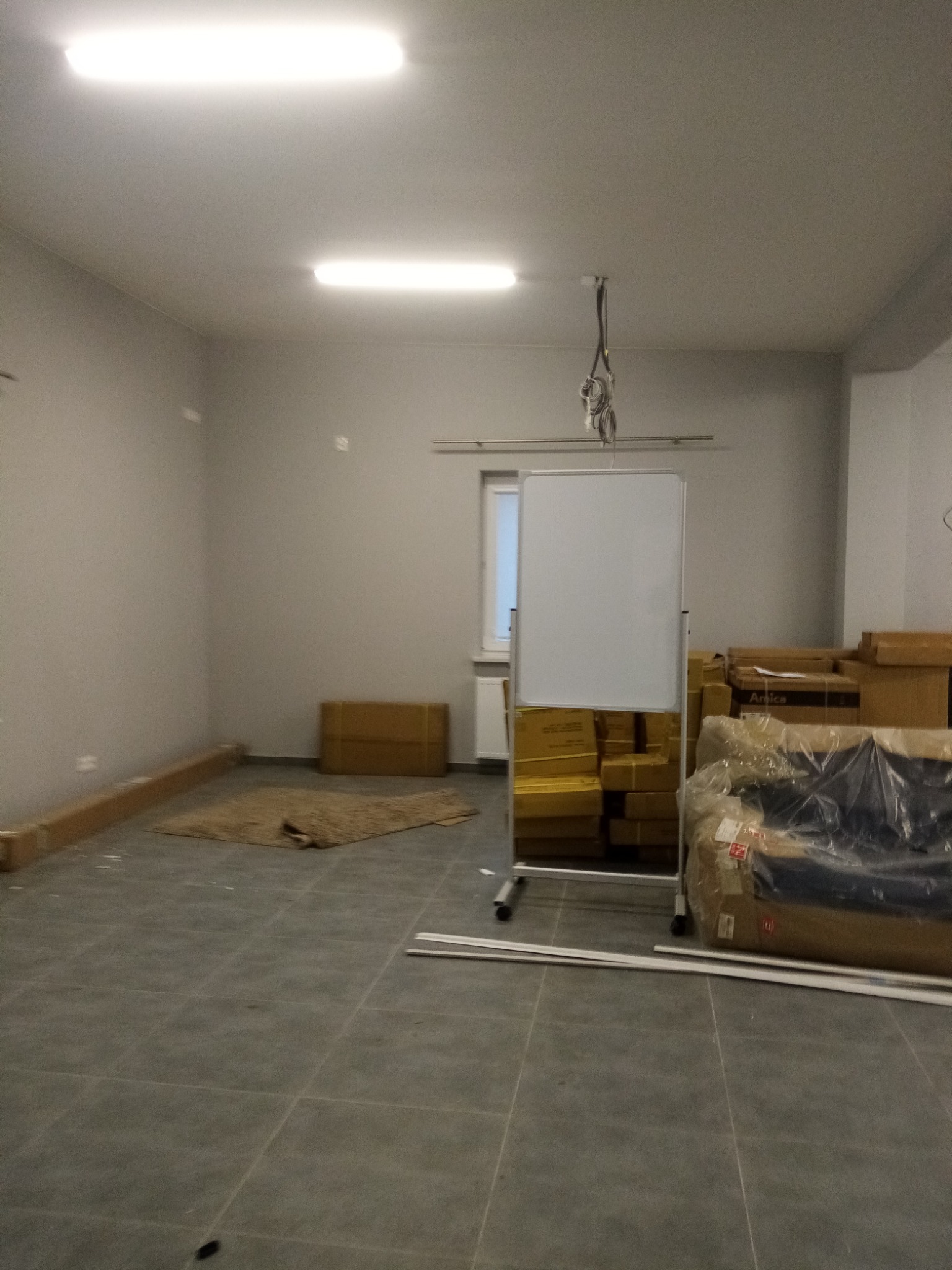 Puste pomieszczenie z szarymi płytkami podłogowymi, przygotowane do prac wykończeniowych, widoczne kartony, folia ochronna i biała tablica na kółkach.